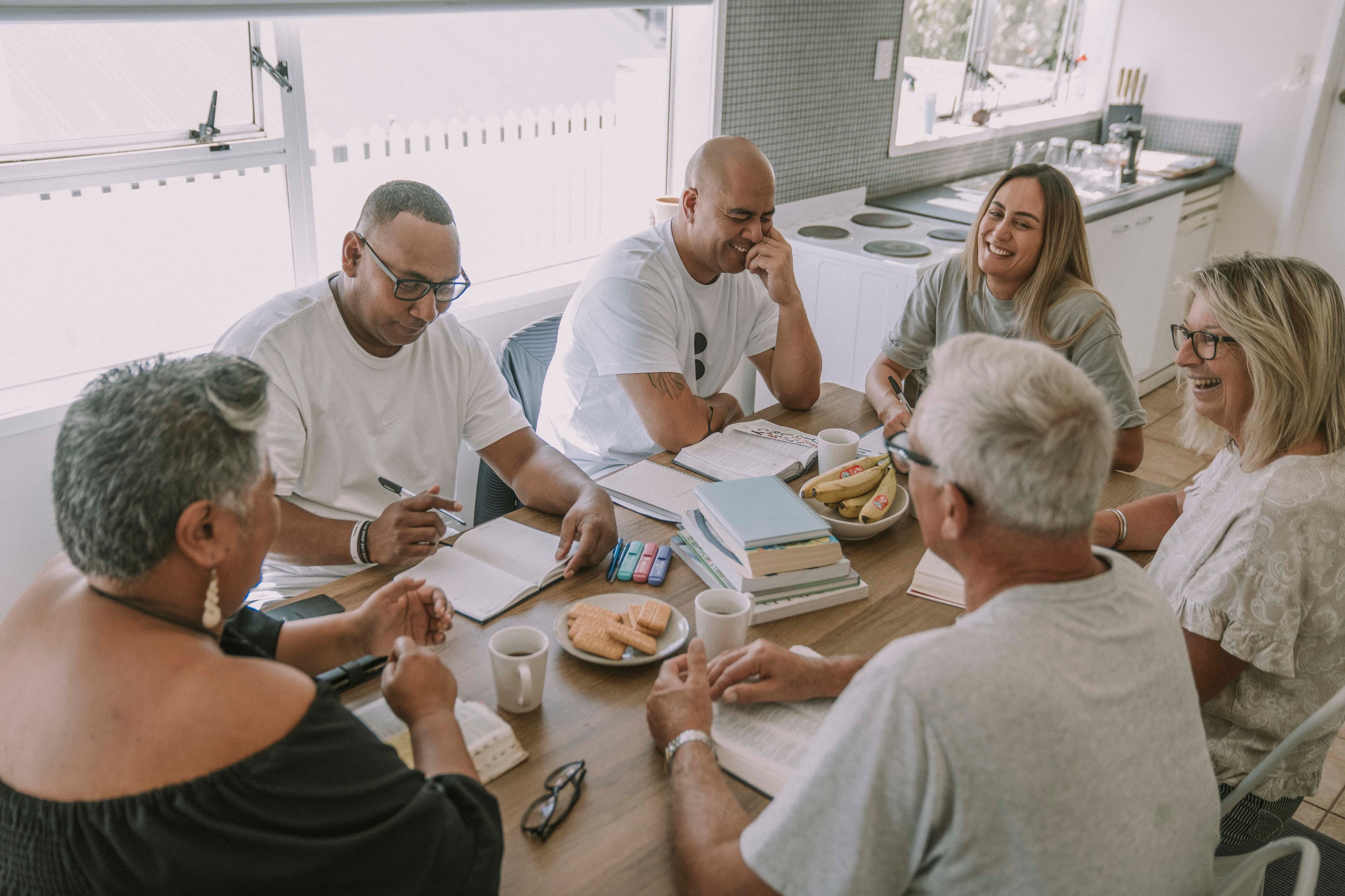 Group of people sitting around an inside table with bibles, notebooks, pens and hot drinks and listening to each other and smiling and laughing