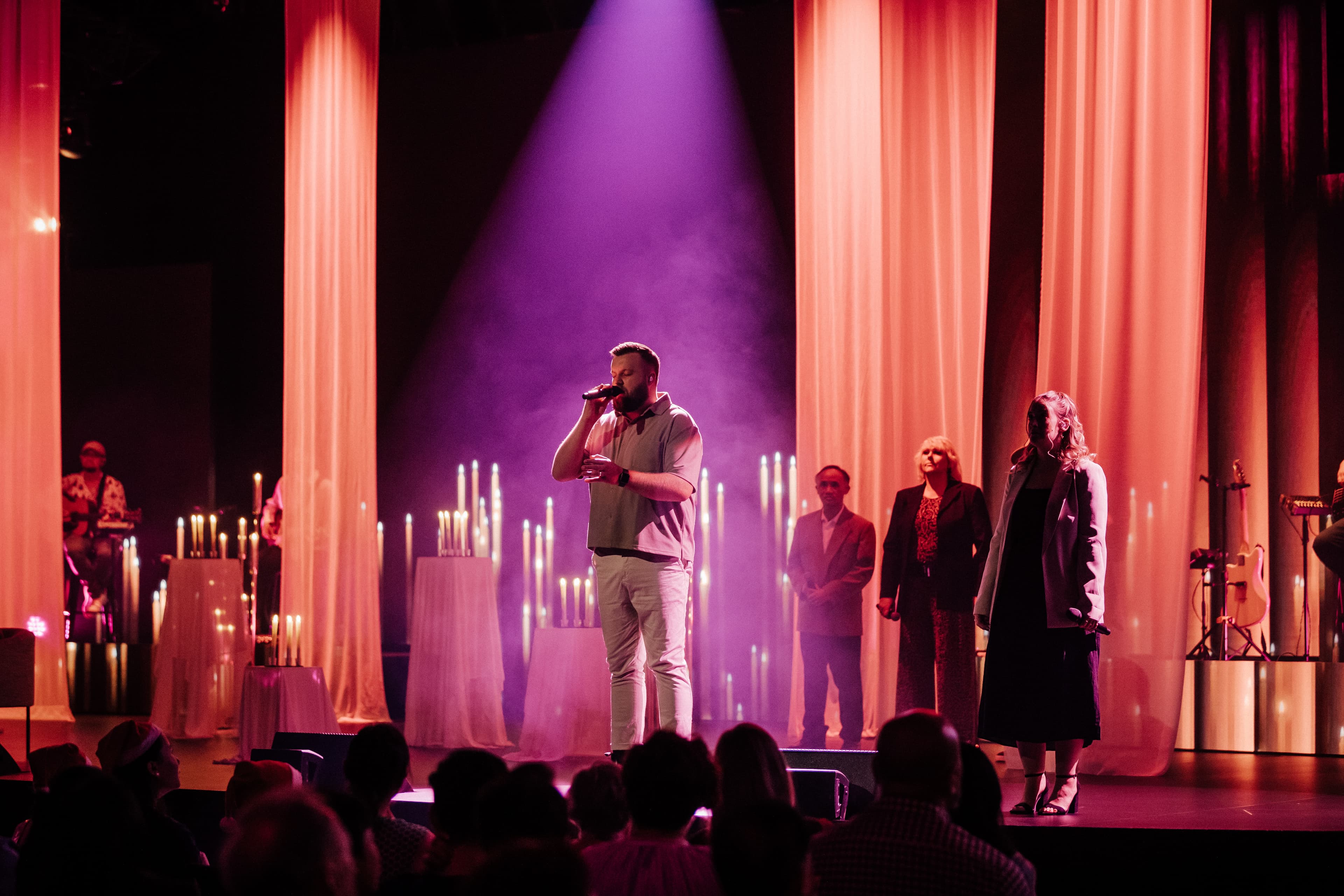 Male singer with backup singers on a pink and orange lit stage with candles on stands in the background