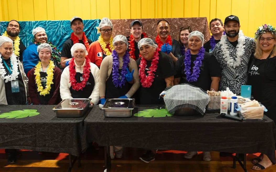 Team of volunteers at a LIFE Community Kitchen in aprons, hairnets, and leis for Samoan Language Week, ready to serve dinner to the diners