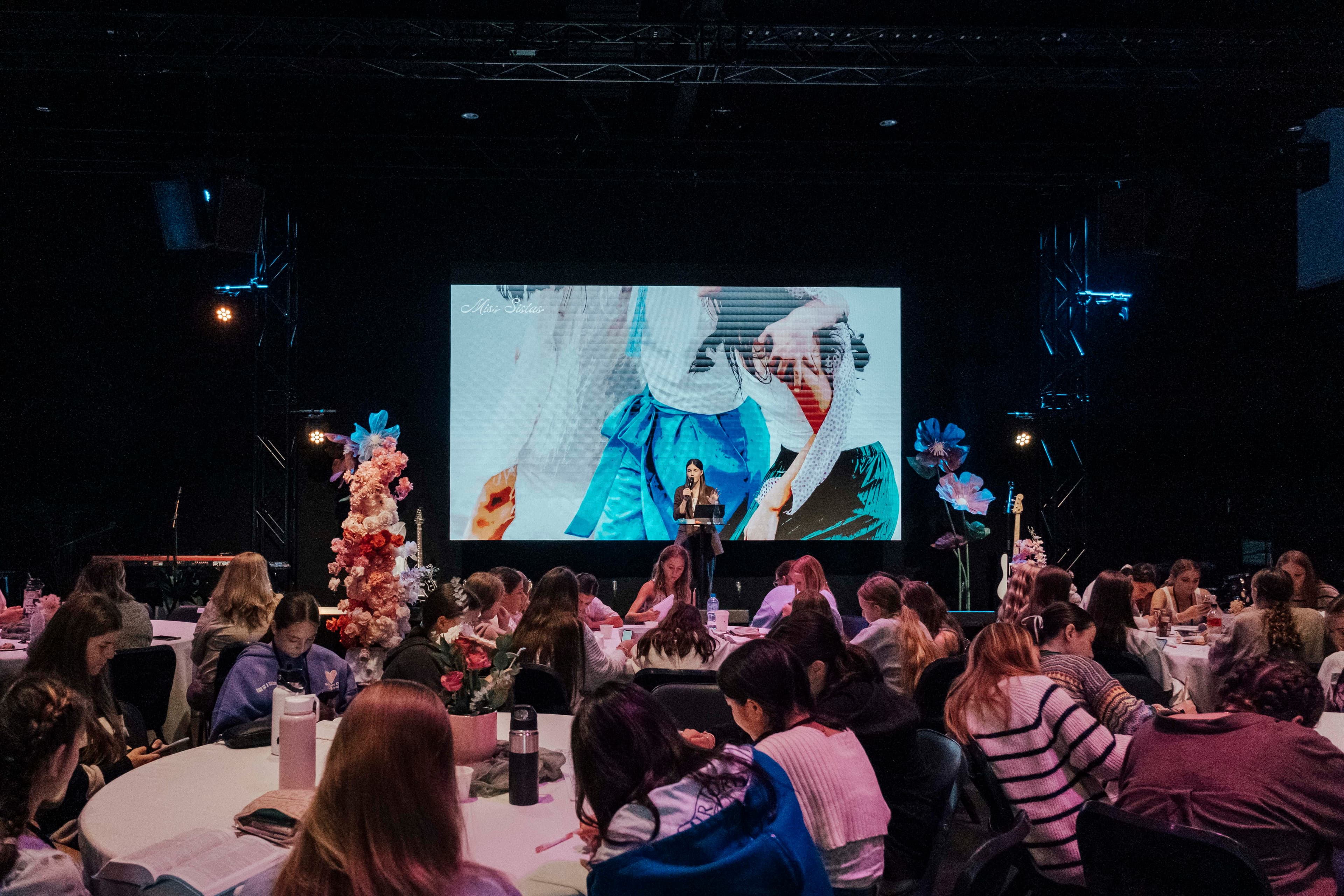 Female pastor on the Chapel stage with a large screen behind her and young women sitting at round tables listening to her speak