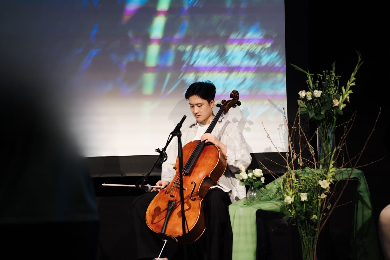 Man playing cello on stage with a floral arrangement on his right