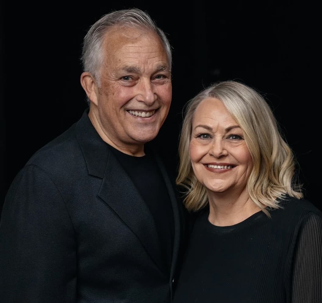 Ps John and Helen Burns smiling at the camera in dark clothing with a black background