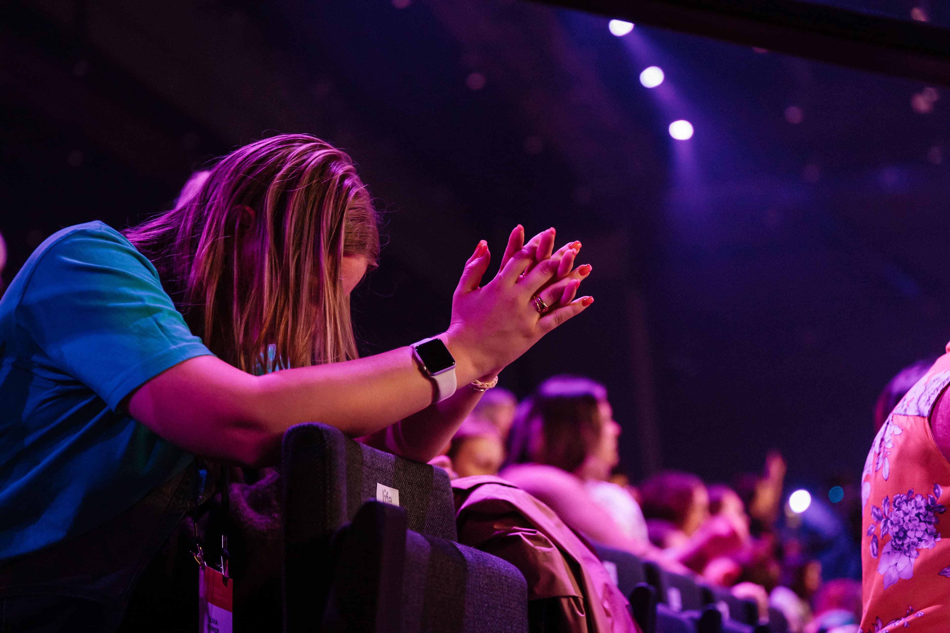 Woman sitting with elbows rested on seat in front of her and hands clasped with head down and covered by her hair in prayer